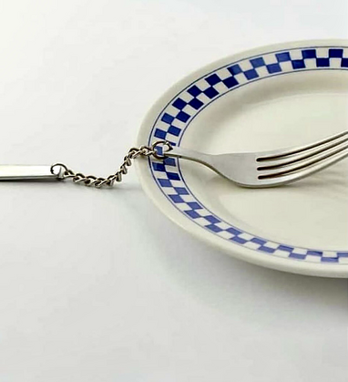 White plate with blue checkered border and a fork lying on top of it where half of the fork is a chain making it impossible to eat with.