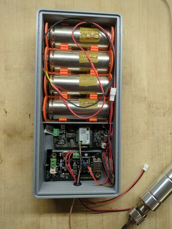 A 3D-printed box with a battery pack taking up about two thirds of the case. The other third contains two PCBs: a RAK Wireless board and a DFRobot solar power management board. There are wires going through a grommet in the side of the case. One of those wires is to a huge antenna, while the others appear to be power connectors.