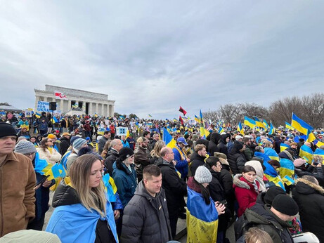 Crowd of demonstrators at a rally, holding Ukrainian flags, near a monument under cloudy skies, showcasing solidarity and support.