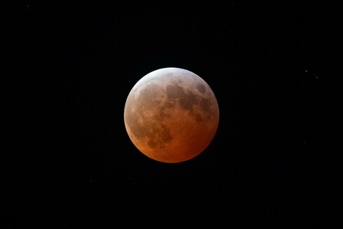 The moon in totality during a total lunar eclipse. A warm orange gradient stretches from the bottom right, a deep orange-red where the shadow is most intense, to an almost white-gray at the top left where the shadow is weakest. The surface of the moon is full or large craters much grayer than the normally bleached gray surface, now all bathed in orange.