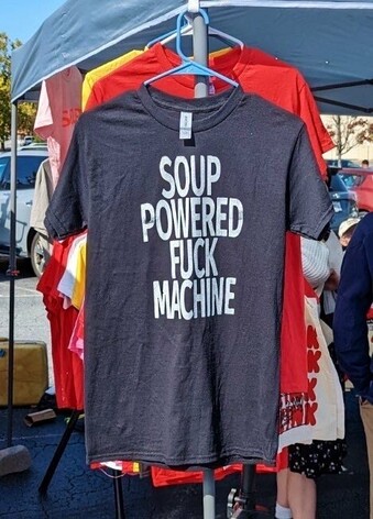 A black t-shirt displayed on a hanger at an outdoor market. The shirt has bold white text that reads:

“SOUP POWERED FUCK MACHINE.”