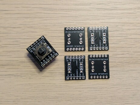 A photo of some small circuit boards on a desk. One has had a small 5-way switch and header pins soldered to it, the other four are plain. They say "@timixretroplays" and "ADA504 breakout" on them and the pins are labeled A, B, C, D, S and Common.