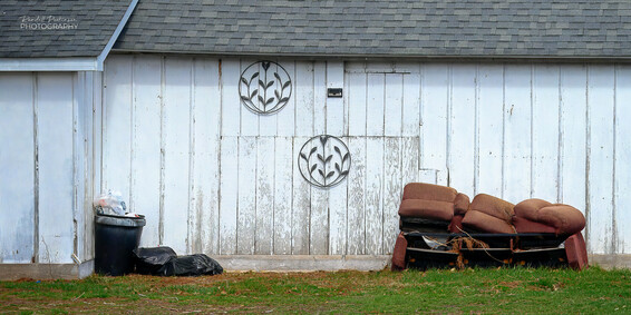 A trash can, two plastic bags of trash, and an old, falling apart sofa site next to a white garage building.