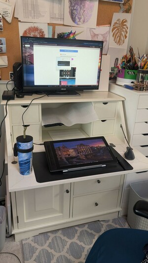 Photo of a white writer's desk with a blue chair on the bottom right. Above the desk are carious foiled prints. To the right is a large spinning caddy of pens and art tools. Below it is a tall bureau of drawers. The desk has a large monitor on top. On the main desk is a large blue tumbler with a straw, a huion drawing tablet and a mouse.