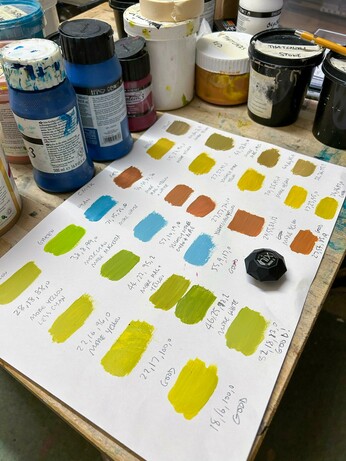 A lot of colour swatches painted onto a sheet of paper with CMYK values written below them. On the paper also sits a Nix colour sensor.