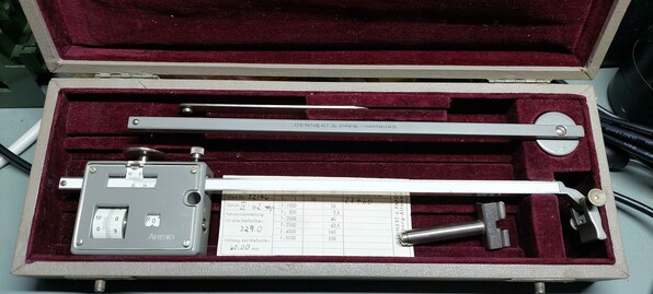 A grey metal tool inside a velvety cushioned box.
The tool has a few scales and wheels on one component. Other pieces may or may not attach to it.