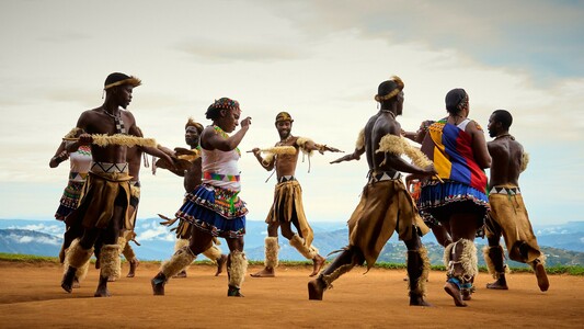 Several dancers perform outdoors, dressed in colorful traditional Zulu attire. The dancers wear beaded skirts and feathered accessories, moving energetically in a circle. The background displays a vast open landscape under a cloudy sky.
