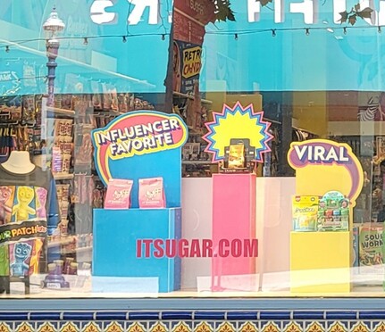 An 'its sugar' candy store window with display of candies referred to as Influencer and Viral, and on top, unlabeled POW! Colorful cardboard cutout.