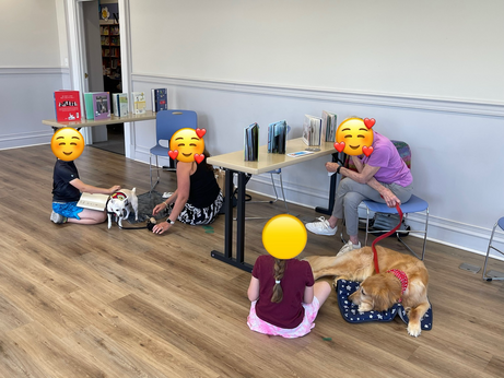 Children reading to dogs under the attentive and caring eyes of the town's neighbors. A golden setter lays lazily down next to a child with a long pony tail listening to her read. Beside this group a young boy pets a tiny chihuahua while holding a book in his lap.
