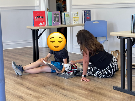 A woman sits on the ground petting her dog while a young boy reads to both of them.