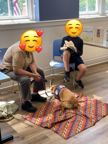 An older boy sits in a chair, flipping through pages of a thick book while reading to a dog laying beside him. An older lady leans forward, attentive and encourage. The doggo is a good boy.