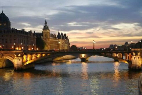 Sunset view of the Seine River in Paris with the Pont du Change bisecting the photo at midpoint and the Conciergerie Castle rises on the left. The structures are shown in the glow of the  pale orange setting sun, muted by a few gray clouds streaking the sky, and several electric lights on the bridge  and reflecting on the river add to the illumination.