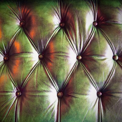 The back of a deep green, modern leather-upholstered, button tufted, chesterfield-styled sofa fills the frame under amber accented lighting.