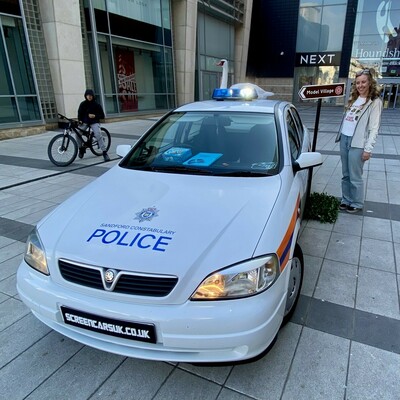 White Vauxhall Astra marked as a "Sandford Constabulary Police" car, parked on a tiled plaza outside a shopping centre. A fake swan is mounted on the roof, referencing the film Hot Fuzz. A sign behind the car points to a “Model Village.”