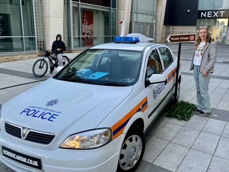 White Vauxhall Astra marked as a "Sandford Constabulary Police" car, parked on a tiled plaza outside a shopping centre. A fake swan is mounted on the roof, referencing the film Hot Fuzz. A sign behind the car points to a “Model Village.”