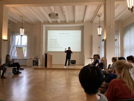 Large seminar room with nice wooden flooring with participants sat on chairs in a circle. On the board, there are some bullet points about International Sign.