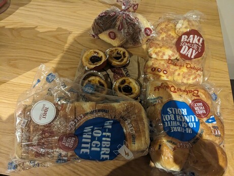 A bunch of bakery items in Bakers Delight bags. It features a loaf of bread, 6 X bread rolls, 6 X mini cheese and bacon rolls, 4 x Portuguese tarts and 4 x scones.