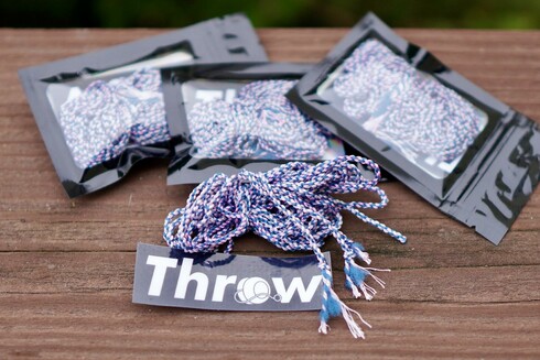 four packs of Trans Pride strings by Throw-Yo, plus a sticker that says "Throw". Each pack contains 5 yo-yo strings and each string has a mix of white, pink, and blue threads. The final strings look similar to the Trans Pride flag colors.