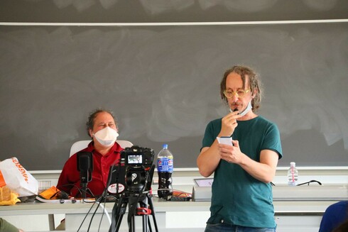 The image depicts an indoor scene, likely a classroom or lecture setting. In the foreground, a person stands holding a piece of paper, wearing a teal t-shirt and glasses, with a microphone clipped to their shirt. They appear to be speaking or presenting. A camera on a tripod is positioned in front of them, suggesting a recording or live broadcast is taking place. The person is wearing a face mask.

In the background, another individual is seated at a desk, wearing a red shirt and a white face mask. They are positioned behind a microphone and a large blackboard, which is blank. On the desk, there is a bottle of Pepsi, a water bottle, and a bag with orange text, possibly a snack or food item. The setting includes a whiteboard and a table, with various items scattered on it, including a yellow cloth and a microphone stand. The overall atmosphere suggests an educational or professional environment.