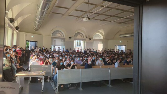 he image depicts a large indoor gathering in a spacious room with high ceilings and arched windows, allowing natural light to fill the space. The room is filled with a crowd of people, mostly young adults, seated in rows of benches. Many individuals are wearing face masks, suggesting a health-conscious environment, possibly due to a pandemic. The attendees are dressed in casual attire, with some carrying backpacks, indicating a student-like atmosphere. The room features a modern design with visible air ducts and ceiling lights. The audience is focused on something off-camera, likely a speaker or presentation. The overall scene conveys a sense of organized assembly, possibly for an educational or informational event.