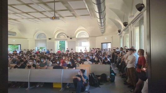 The image depicts a large indoor gathering in a spacious room with high ceilings and arched windows, allowing natural light to fill the space. The room is filled with a crowd of people, mostly young adults, seated in rows of benches. Many individuals are wearing face masks, suggesting a health-conscious environment, possibly due to a pandemic. The attendees are focused on something in front of them, likely a speaker or presentation. Some people are standing in the aisle, possibly waiting for their turn to sit or to get a better view. The room has a formal atmosphere, with a sense of order and attentiveness among the crowd. The text "AMENTE CHIUSO" is visible on a sign, indicating that the room is closed or restricted. The overall scene suggests a public event, possibly educational or informational, held in a setting that accommodates a large number of participants.
