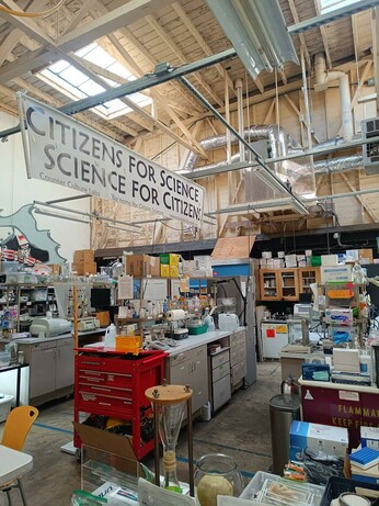 A community lab in a warehouse with rows of lab benches and mostly vintage lab equipment. A large banner reads "citizens for science, science for citizens"