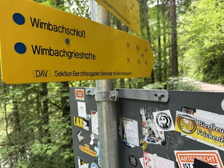 Ein gelbes Schild im Wald, mit der Aufschrift „Wimbachschloß
Wimbachgrieshütte
DAV Sektion Berchtesgaden, Nationalpark Berchtesgaden“

Darunter die Rückseite eines anderen Schildes, auf dem ein Sticker „Abgelenkt und trotzdem da – ADHS Antifa“ klebt.