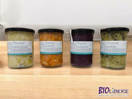 Cuatro frascos de vidrio con tapas negras se alinean en una superficie de madera clara. Cada frasco contiene un tipo diferente de chucrut fermentado: chucrut tradicional, baechu kimchi, chucrut lombarda y chucrut eneldo. Las etiquetas de cada frasco indican el nombre y tipo de chucrut en español. El fondo muestra el logo de "BIOLIBERE supermercado cooperativo".