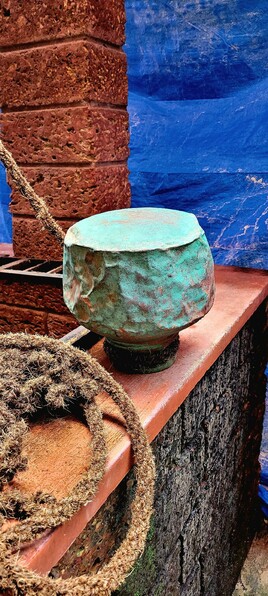 A heavily rented and oxidized-green copper pot placed upside down on the wall of a well. Red walls. A thick drawing rope in the left foreground. Blue tarp in the background.