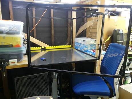 A box made of black aluminum 20 mm t-slot rails.  The sides are empty and the bottom is an aluminum sheet.  The front door is swung out in front of a blue chair.  The entire aluminum frame is on a custom wooden desk framed from 2x4 beams with bright LED overhead lighting.
