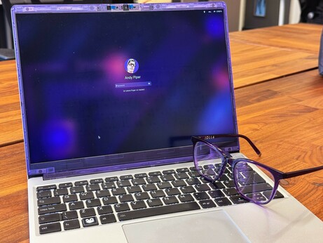 A Framework laptop with a translucent purple bezel sits on a wooden table, showing a login screen for the user "Andy Piper." A pair of purple-framed Iolla spectacles rests on the keyboard.