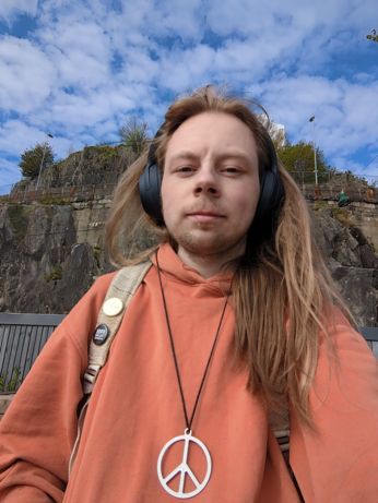 The image features a person wearing a large, round silver peace pendant necklace, an orange hoodie, and black headphones. The individual has long, light brown hair and is standing outdoors with a rocky hillside and a blue sky with scattered clouds in the background. The person is also wearing a beige backpack with a visible button that reads "FREE JULIAN ASSANGE" The setting is a public area with a metal fence and street lamps visible in the background.