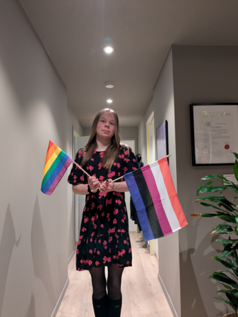 The image shows a person standing in a hallway, holding two flags. The individual is wearing a black dress with a floral pattern in pink, paired with black tights and knee-high black socks. The person is holding a rainbow pride flag on the left and a flag with a gender-fluid flag in the right. The hallway has light-colored walls and a wooden floor, with recessed ceiling lights illuminating the space. On the right side of the image, there is a framed certificate on the wall and a green plant. The overall setting appears to be a residential interior.
