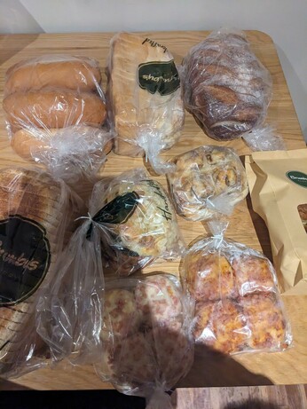 A table filled with bakery goods, namely. 3 X loaves (brown, white, sourdough) in seperate bags, 4 X croissant in a paper bag, 6 hot dog rolls, 4 X mini cheese and bacon, 4 X mini pizza, 4 X cheesy mites and in a final bag an almond croissant, escargot and blueberry danish.