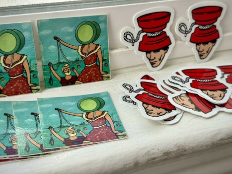 Stacks of stickers in two different styles. One is a yo-yo themed parody of the album cover for “In The Aeroplane O er The Sea” by Neutral Milk Hotel. It shows an old timey drawing of people swimming in a lake and a woman sitting by the edge of the water, but she has a yo-yo for head in this version. 
The other sticker is a yo-yo themed parody of DEVO’s old logo. It shows a man in a red hat, but in this version the hat is actually a giant yoyo.