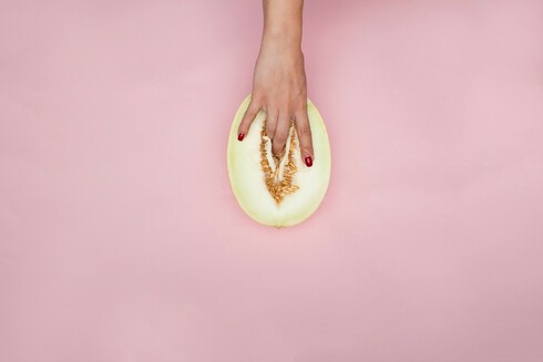 Image of someone dipping fingers down into a sliced white melon between orange seeds, as if it were a vulva, on a pink background. Photo by Deon Black.