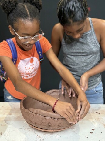 Two young brown-skinned girls are working together on creating the base of an udu. One girl, with her hair in two Afro puffs, wearing glasses and an orange shirt, is smiling as she shapes the clay, while the other girl, with her hair in a ponytail, in a gray tank top, focuses on the task. Their hands are engaged, attaching a coil to the bowl-shaped foundation below.