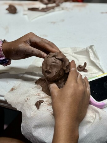 A pair of brown hands is shaping a figure out of brown clay on a white table. The hands appear to be working with the clay, which is messy and smudged with dirt. A phone is visible at the edge of the table.