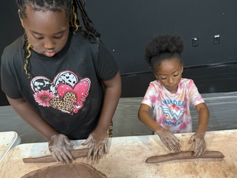 Two children are engaged in a clay modeling activity. One child wears a black t-shirt with hearts and focuses on rolling a piece of clay, while the other child, dressed in a tie-dye "Wonder Woman" shirt, is also rolling clay.