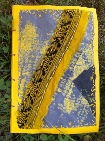 The cover of a yellow-blueish art book showing a diagonale of a silk strip on a maize cobble print. The book cover is made with sun-yellow cardboard. The cover itself: an abstract print collage of blue coloured krafft paper with a yellow print of maize cobbles and intuitive yellow embroidery at the borders. A stripe of yellow saree silk waste with a beautiful black pattern is sewed on the cardboard with white embroidery.