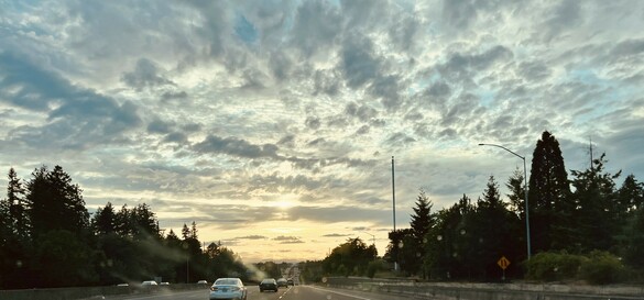 Looking through my windshield at a long road ahead, lined on each side with lots of trees. The sky is filled with billowy clouds. It’s a gradient color, beginning with a light blue gray overhead, continuing to a light gold where it meets the earth. The bright sun is trying to peek through a layer of clouds near the horizon.