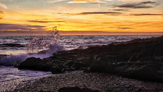 A dramatic sunrise over a rocky coastline in the east of Ireland. The sky is ablaze with vibrant orange and gold hues, with thin cloud streaks and faint contrails visible. In the foreground, dark, rugged rocks line the shore, with a powerful wave crashing against them, creating a spectacular spray of white foam. The ocean stretches into the distance, appearing dark and choppy under the colourful sky, with a line of wind turbines silhouetted against the horizon. The overall mood is powerful and awe-inspiring.