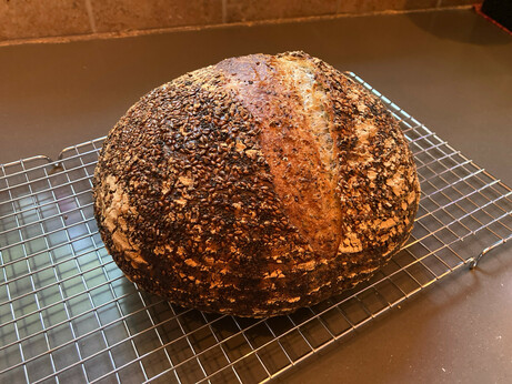 A heavily-seeded loaf of bread sits on a silver cooling rack. It is very dark brown, and the crust looks very crispy.
