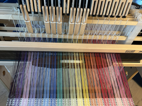 A work in progress on a hand loom showing a wooden boat shuttle midway through a pass through a warp in a rainbow of colours