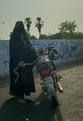 A woman in full hijab on the side of the road, standing next to her motorbike, one hand on the saddle, facing the camera. Palm trees peek out over a wall with blue Islamic calligraphy in the background.