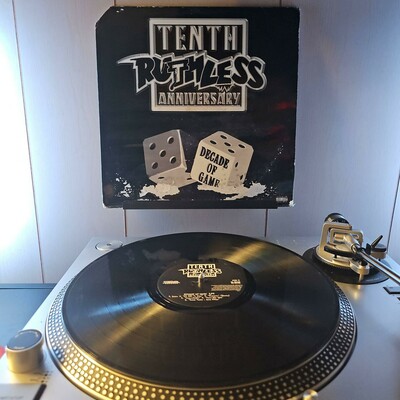 A vinyl record sits on a turntable. Behind the turntable, a vinyl album outer sleeve is displayed. The front cover shows a pair of dice, with one that says "Decade of Game". Text above says Tenth Ruthless Anniversary.  There is damage to the album cover