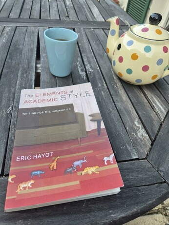 Sur une table en bois, le bouquin est posé à côté d'une tasse bleue et d'une théière ocre à pois colorés.