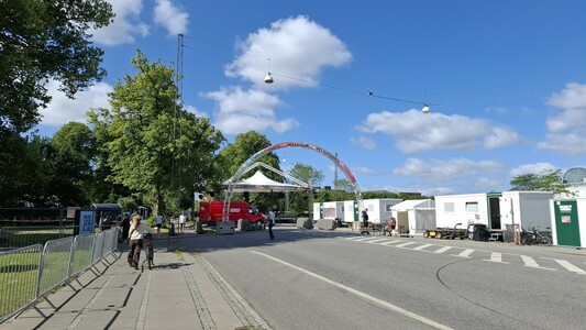 Earlier today, people seeing up a European Street Party arch and other public event paraphernalia on a road by city hall that is closed to cars. People are walking and cycling around as normal. It is sunny amd windy. The sky is blue with white clouds.