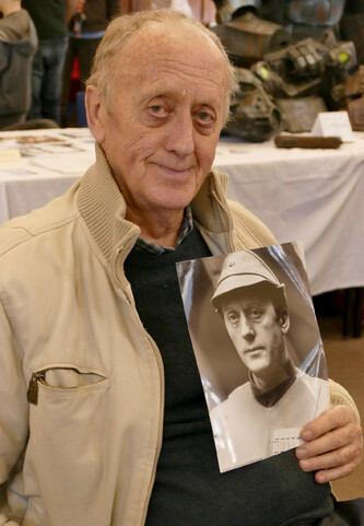 A picture of Kenneth Colley sitting at a convention, holding a photograph of himself in Star Wars.