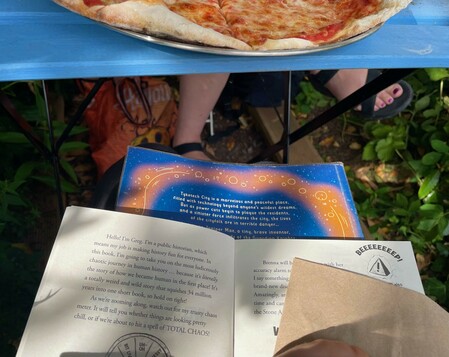 Beneath a blue outdoor picnic table with pizza on top is the paperback book spread wide open for reading.